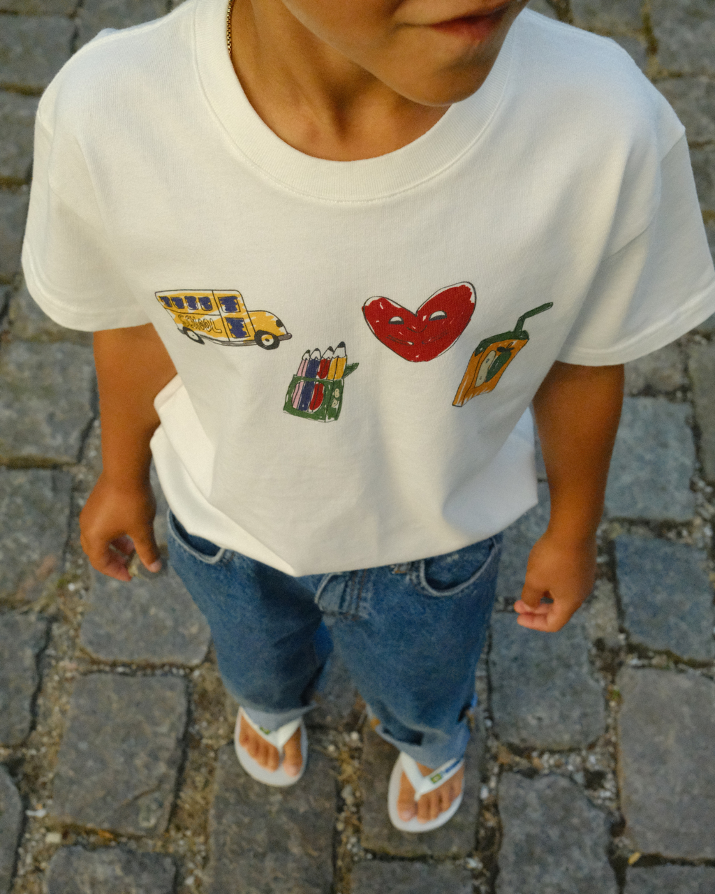 A child wearing a white short-sleeve T-shirt with colorful graphic illustrations of a yellow school bus, a red heart, a pencil box, and a pepperoni slice, paired with blue jeans and white sandals, standing on a cobblestone street.