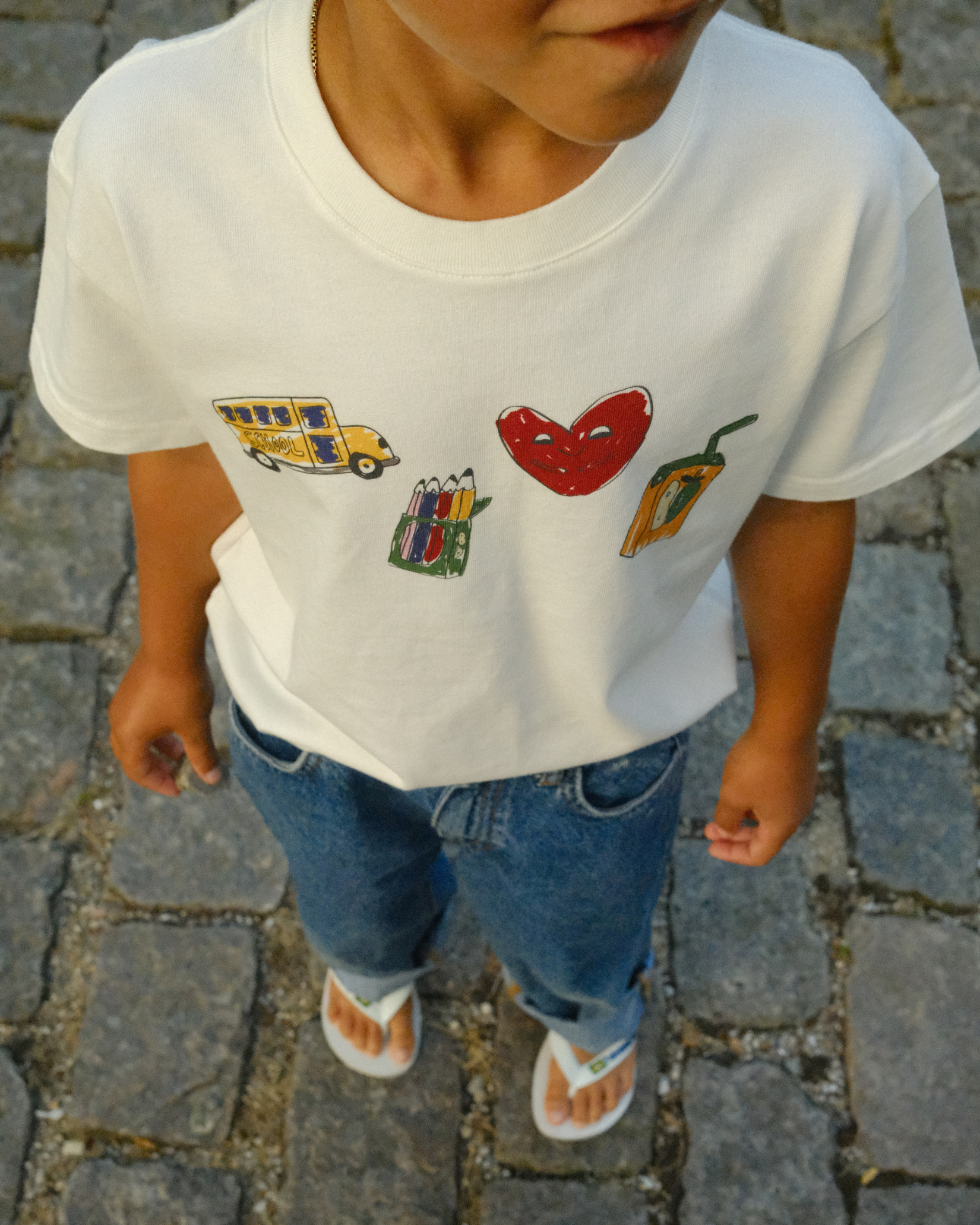 A child wearing a white short-sleeve T-shirt with colorful graphic illustrations of a yellow school bus, a red heart, a pencil box, and a pepperoni slice, paired with blue jeans and white sandals, standing on a cobblestone street.